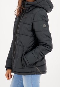 Vrouw in een zwarte gewatteerde jas met een capuchon, hartvormig logo op de borst en mouw, en lichtblauwe jeans, staand met één hand in de zak.