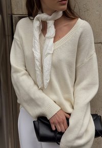 Cremefarbener gestrickter Pullover mit V-Ausschnitt und oversized Silhouette. Kombiniert mit einem strukturierten weißen Schal und einer schwarzen, sanft gestalteten Clutch.