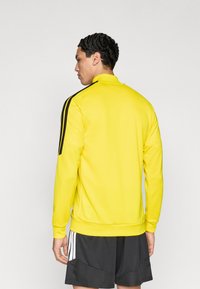 Κίτρινο αθλητικό φούτερ με ψηλό γιακά, μακριά μανίκια και μαύρες ρίγες στους ώμους. Λεπτή υφή, σε συνδυασμό με μαύρο σορτς.