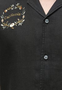 Μαύρο πουκάμισο με κουμπιά με κεντητό το «California» με κίτρινη κλωστή μέσα σε ένα λουλουδένιο στεφάνι στο αριστερό στήθος.