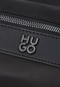 Sort stofpose med en lynlås og en glat læderpatch prægede med "HUGO" i sølv. Teksturen er subtil med et elegant design.