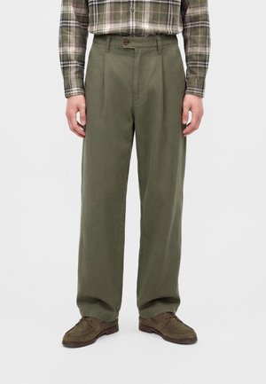 Mann trägt olivgrüne plissierte Hose, braune Wildleder-Schnürschuhe und ein grünes kariertes Langarmhemd mit heruntergelassenen Ärmeln.