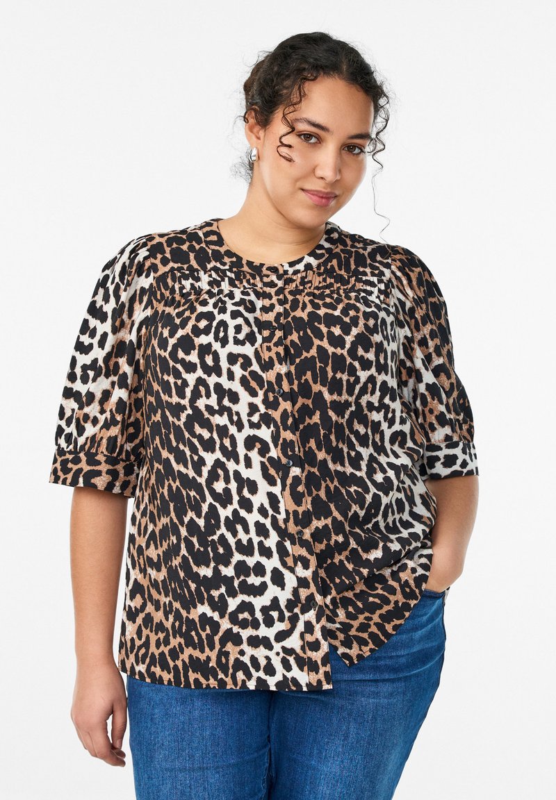 Kučeravá žena s striebornými náušnicami, v blúzke s leopardím vzorom a nafúknutými rukávmi a modrých džínsoch, stojaca s jednou rukou vo vrecku.