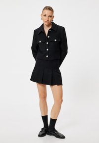 Zwarte tweed blazer met twee vo pockets en grote knopen, gecombineerd met een geplooide zwarte mini rok. Aangevuld met zwarte sokken en loafers.