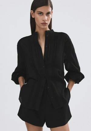 Femeie cu păr lung și închis la culoare, purtând o cămașă neagră cu nasturi și mânecile suflecate și pantaloni scurți negri, stând cu mâinile în buzunare.