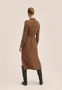 Robe marron avec un motif ondulé en orange et crème, manches longues, taille ceinturée, longueur mi-mollet, fentes latérales, associée à des bottes noires montant jusqu'au genou.