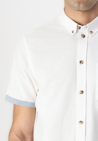 Korte mouwen, wit overhemd met knopen en een kraag, met lichtblauwe manchetten. Heeft bruine knopen en een borstzak. Katoenen stoftextuur.