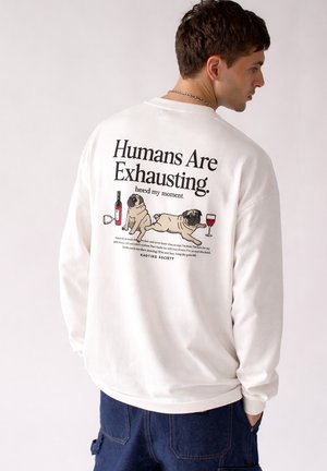 Junger Mann trägt weißen Pullover mit Mops-Abbildungen, Weinflasche, Glas und dem Text „Menschen sind anstrengend“ auf dem Rücken.