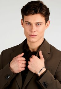 Blazer marrone con finitura opaca, dettagli con bottoni sulle maniche, indossato sopra una camicia scura, modello che indossa un bracciale in argento.