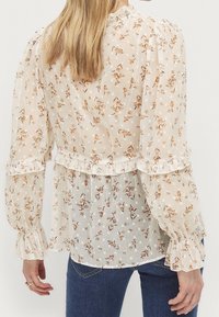 Blusa floreale in tessuto leggero e semitrasparente, con una base crema decorata con fiori marroni e arancioni, polsini arricciati e vita aderente.