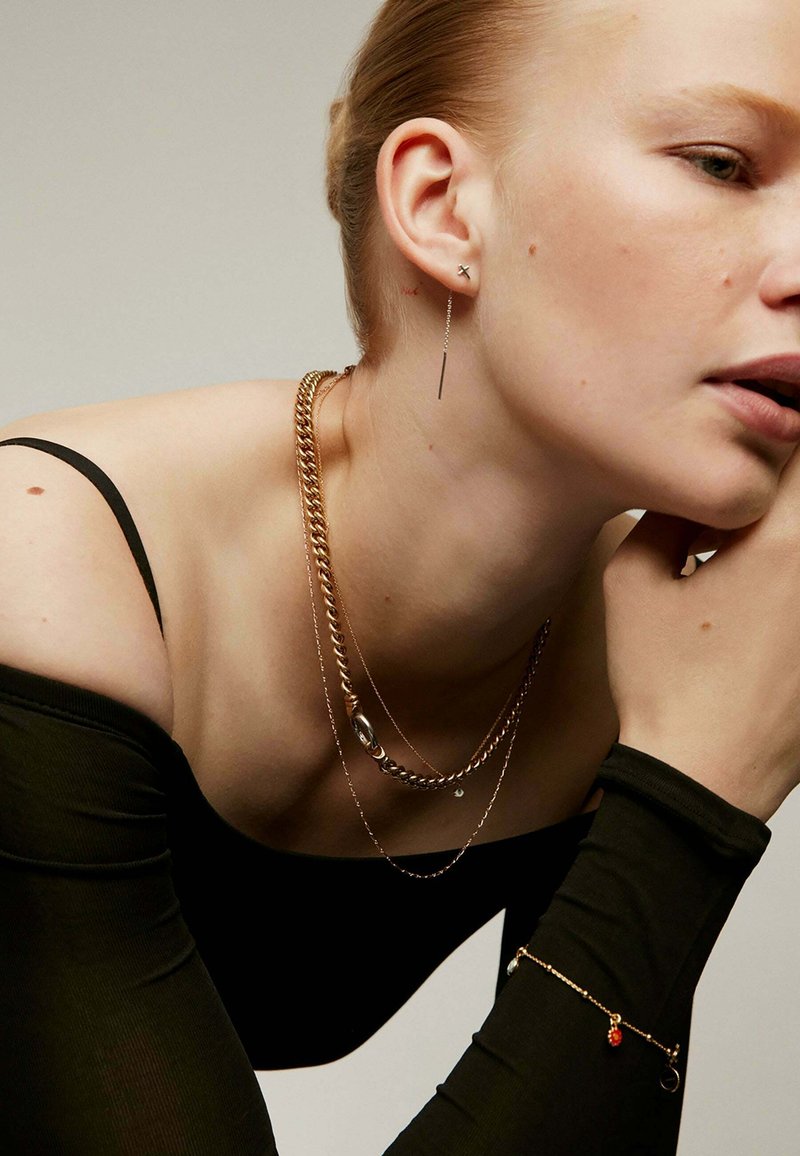 Collares en tonos dorados en capas y un pendiente colgante con un diseño lineal, combinados con una blusa negra de hombros al descubierto y una pulsera detallada.