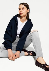 Haut à capuche zippé bleu marine avec une coupe spacieuse, porté par-dessus un t-shirt blanc. Jean déchiré gris clair et chaussures plates brun foncé avec des accents métalliques.