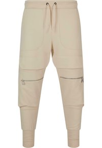 Beige joggebukser med snor i livet, to frontlommer, glidelåslommer på siden, ribbestrikkede mansjetter, og en glatt bomullsblanding.