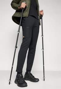 Zwarte wandelbroeken met ritszakken op de zijkant, gecombineerd met zwarte wandelschoenen. De outfit wordt aangevuld met een olijfgroen jack.