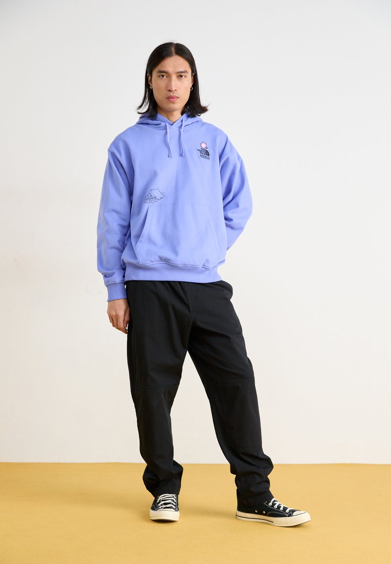 Light purple hoodie with a front pocket and graphic design, paired with black trousers and black sneakers. Simple, relaxed fit.