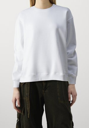 Henkilö, joka pukeutuneena yksiväriseen valkoiseen pitkähihainen pyöreäkauluspaitaan ja tummanvihreisiin cargo-housuihin, seisoo vaaleaa taustaa vasten.