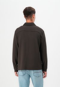 Camisa de manga larga marrón oscuro con cuello, con una textura suave y un diseño de yugo. Combinada con jeans de mezclilla azul claro.