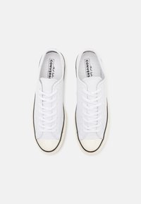 Bílé plátěné tenisky s texturovaným povrchem, kulatou špičkou, černými gumovými akcenty a bílými šňůrkami. Logo viditelné na stélkách.