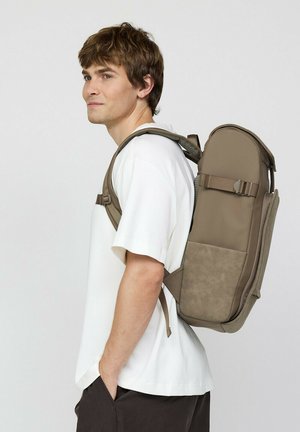 Junger Mann trägt ein weißes T-Shirt und dunkle Hose und trägt einen großen taupefarbenen Rucksack mit mehreren Riemen vor einem schlichten Hintergrund.