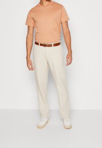 Ljusbrun bomullströja, beige linnebyxor, brunt läderbälte och vita sneakers med ljusbruna sulor, stående på en enfärgad bakgrund.