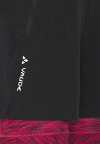 Černé sportovní šortky s texturovaným povrchem, vybavené boční kapsou na zip a bílým logem "Vaude". Pod nimi jsou viditelné růžové legíny se vzorem.