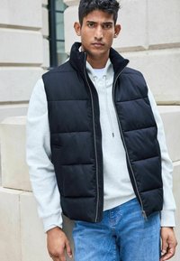 Zwarte puffer vest met hoge kraag, ritssluiting aan de voorkant en zijzakken, gedragen over een lichtgrijze hoodie, gestyled met blauwe jeans.