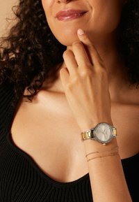 Silberne und goldene Uhr mit rundem Ziffernblatt, Kristallakzenten und einem metallischen Armband, am Handgelenk positioniert. Schwarzes, strukturiertes Oberteil sichtbar.