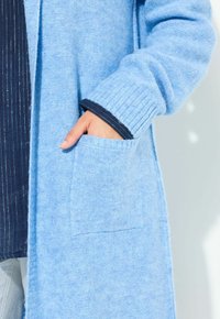 Ljusblå cardigan gjord av mjukt, texturerat tyg, med en ficka i patchstil och ribbade ärmslut med en kontrasterande marinblå kant.