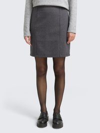 Donna che indossa una gonna grigia a quadretti, collant neri trasparenti, mocassini neri e un maglione a maniche lunghe grigio chiaro, in piedi su uno sfondo neutro.