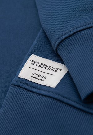 Niebieska tkanina złożona, z białą przyszytą metką z napisem „Twoim jedynym ograniczeniem jest umysł” oraz nazwą marki „OMBRE od 1985.”