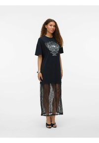 Femme portant une robe tee-shirt noire oversize avec un texte graphique, une superposition de jupe en dentelle noire transparente, des talons ouverts noirs, et un bracelet argenté, se tenant devant un fond blanc.