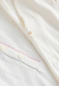 Tissu blanc doux avec de petits motifs floraux, texture délicate en broderie anglaise, et bord boutonnière avec une couture rose en forme d'écailles.