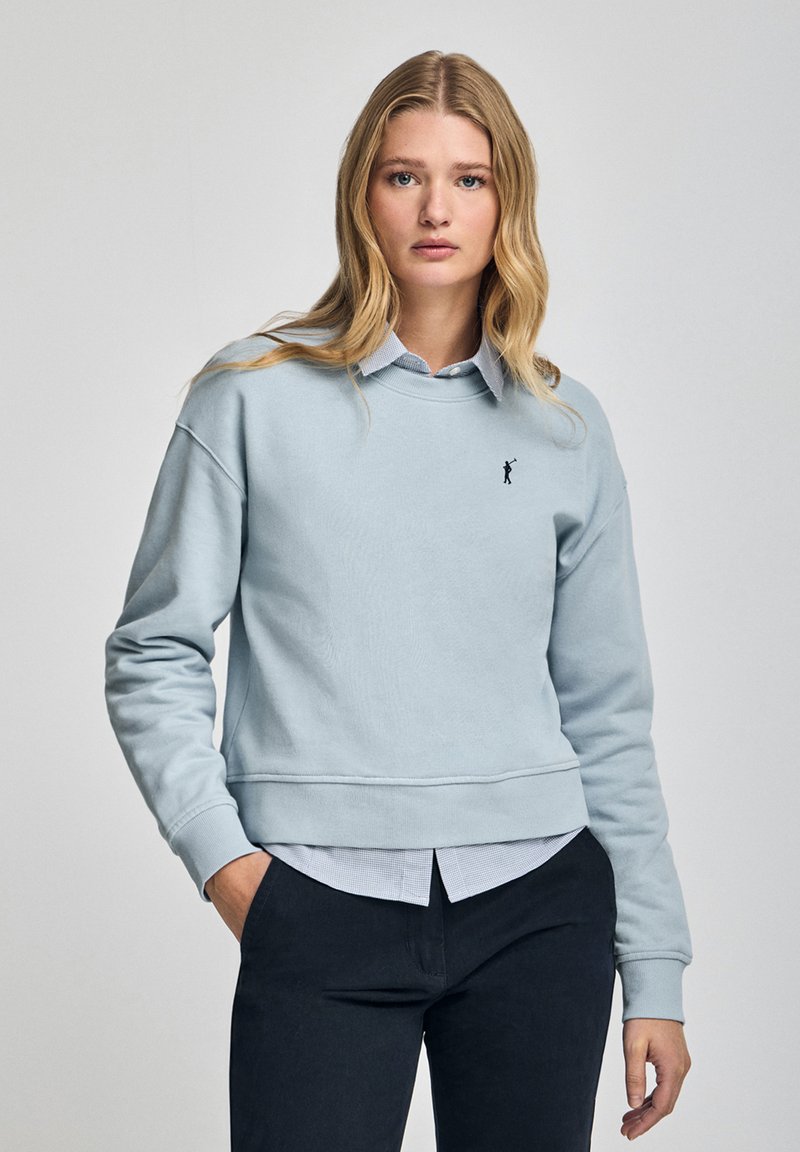 Ljusblå sweatshirt med rund halsringning, ribbade ärmslut och nederkant. Har en broderad logotyp på bröstet och är lager på lager med en skjorta under.