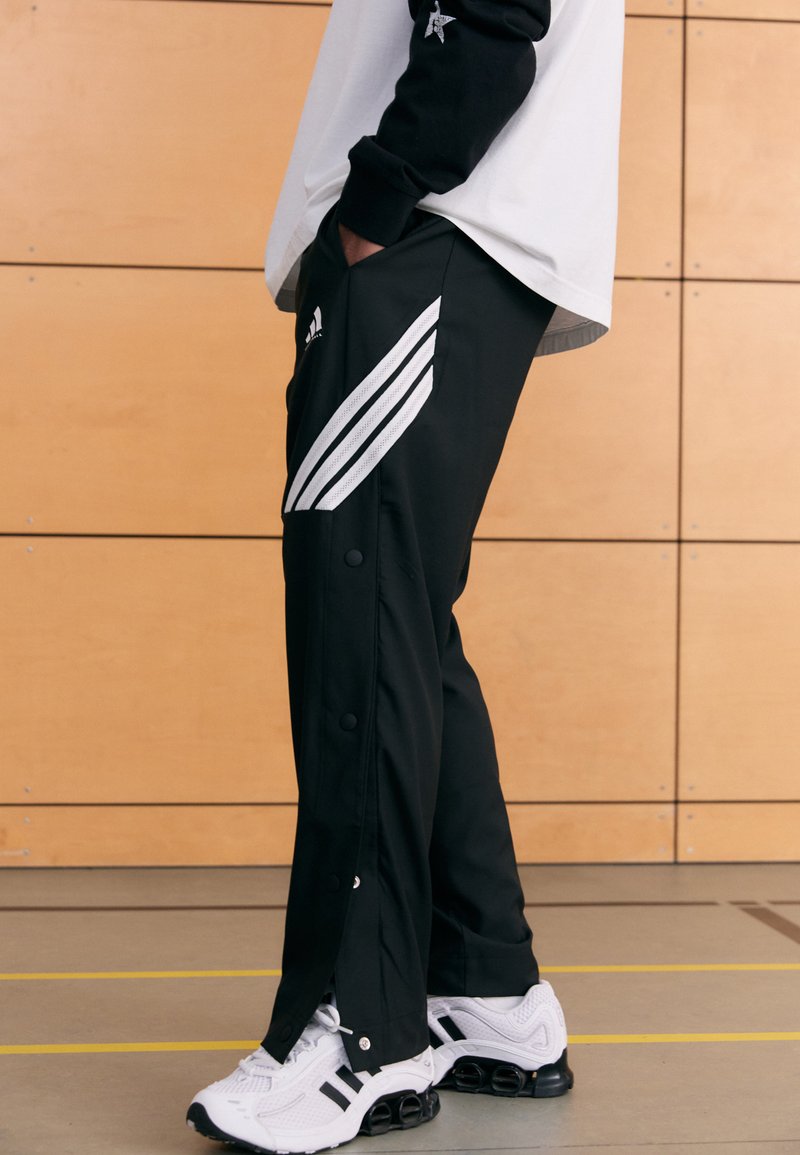 Black athletic pants with white stripe patterns, snap-buttons along the sides, paired with white sneakers featuring black accents.