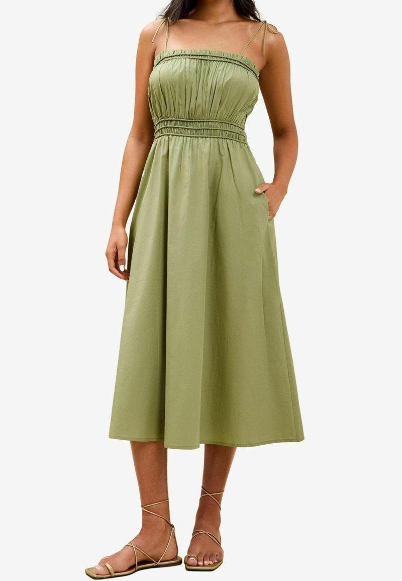 Femme portant une robe midi sans manches vert olive avec un corsage froncé et de fines bretelles, associée à des sandales plates à brides.