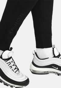Joggers neri con logo "TECH PACK" grigio, abbinati a sneaker Nike Air Max 97 bianco e nero con design a strati e unità d'aria visibile.