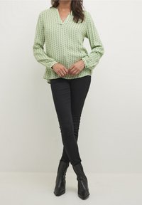 Mujer vestida con una blusa de manga larga de color verde claro y estampada, pantalones skinny negros y botines de tacón puntiagudo negros, de pie con las piernas cruzadas.