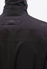 Must jakk kõrge kraega, millel on tagaküljel tikitud brändisilt "HUGO" ning sileda tekstuuriga kangas. Minimalistlik disain.