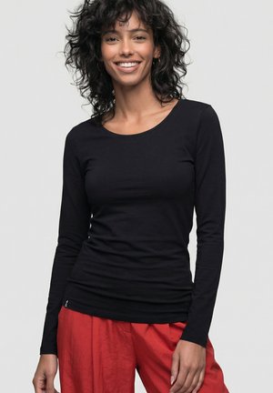 Lächelnde Frau mit lockigem Haar, die ein schwarzes Langarmshirt und lockere rote Hosen trägt, vor einem schlichten hellen Hintergrund stehend.