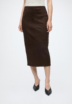 Femme portant une jupe midi marron foncé ajustée et des mules noires à talons pointus, debout devant un fond blanc uni.