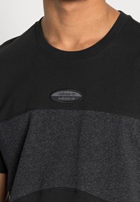 Detailní záběr na osobu v černém a tmavě šedém tričku Adidas s oválným logem Adidas na hrudi.