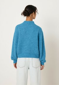 Tamsiaplaukių garbanotų plaukų moteris su bun užsukta, vilkinti mėlyną dryžuotą megztinį ir šviesiai mėlynus džinsus, atsisukusi nugarą prieš paprastą šviesią fono.