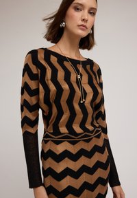 Braun und schwarzer Pullover mit Zickzackmuster, langen Ärmeln und einem Bootsausschnitt. Strukturiertes Strickmaterial, figurbetonte Form und Glanz auf den Ärmeln.