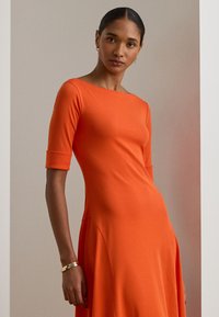 Oranžs ceļgala garuma kleita, izgatavota no mīksta auduma. Tam ir īsas piedurknes un laivu formas apkakle, un tas ir ar plandošu A formas siluetu. Akcentēta ar šuvju detaļām.