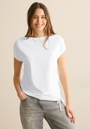 Lächelnde Frau mit langen Haaren trägt ein weißes kurzärmeliges Oberteil mit seitlicher Schleife, graue Jeans, steht mit einer Hand in der Tasche vor einem beigen Hintergrund.