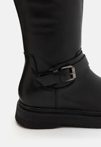 Bota de cuero negro hasta la rodilla con punta redonda, superficie texturizada, detalle de correa ajustable con una hebilla metálica y suela plana de goma.