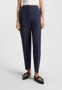 Navy tailored trousers made of a smooth fabric, featuring a front crease, high waist, and side pockets. Paired with black flats.