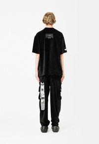 Camiseta de gran tamaño de pana negra con tejido texturizado, que presenta un bolsillo en la parte posterior. Combinada con pantalones cargo negros y calzado negro robusto.