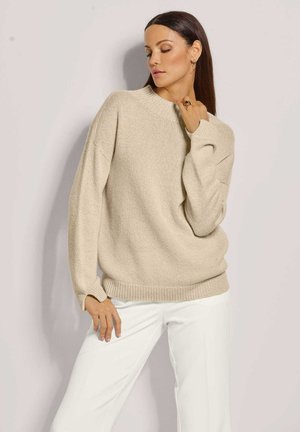 Beige gestrickter Pullover mit hohem Rundhalsausschnitt, überschnittenen Schultern und gerippten Bündchen. Kombiniert mit weißen weiter geschnittenen Hosen.