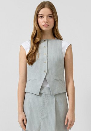 Młoda kobieta o długich włosach, ubrana w jasnoszary, prążkowany, bezrękawnik i dopasowaną spódnicę, na białej bluzce z krótkim rękawem, stojąca na jednolitym tle.